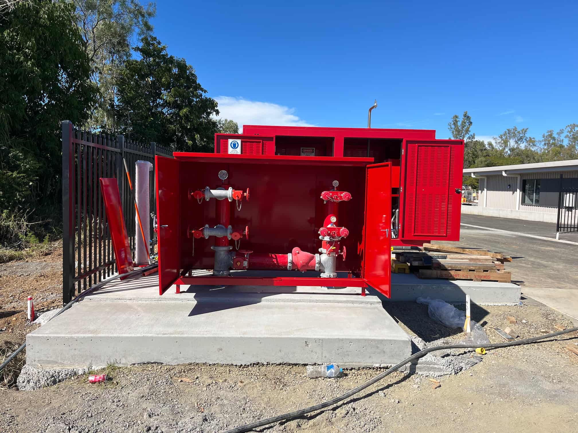 A red fire pump unit installed on a concrete slab with its doors open, showing various valves and piping. The surrounding area is under construction with pallets, pipes, and a fence in the background.